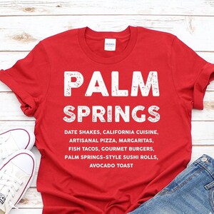 May include: A red t-shirt with the words "PALM SPRINGS" in large white letters. Below, a list of food items is printed. The shirt is paired with denim shorts and white sneakers, suggesting a casual, vacation-themed outfit.