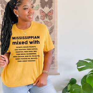Pu&ograve; includere: T-shirt giallo senape con la scritta "MISSISSIPPIAN mixed with" seguita da un elenco di cibi del Sud. L'elenco include Po'boys, pesce gatto fritto e torta di patate dolci. La maglietta &egrave; indossata da una persona.