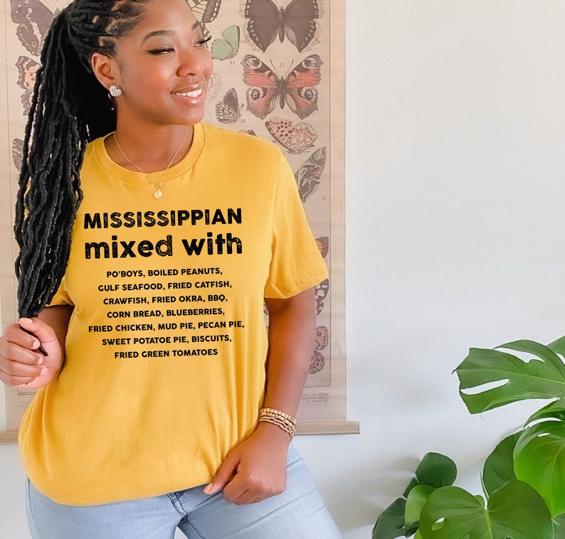 Pu&ograve; includere: T-shirt giallo senape con la scritta "MISSISSIPPIAN mixed with" seguita da un elenco di cibi del Sud. L'elenco include Po'boys, pesce gatto fritto e torta di patate dolci. La maglietta &egrave; indossata da una persona.