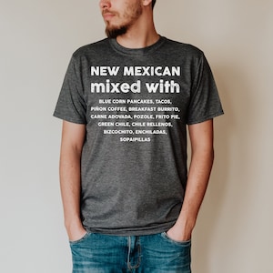 Pu&ograve; includere: Una maglietta grigio scuro con la scritta "NEW MEXICAN mixed with" e un elenco di cibi del New Mexico in bianco. L'elenco include pancake di mais blu, tacos, caff&egrave; pi&ntilde;on e altro. La maglietta &egrave; indossata da una persona.