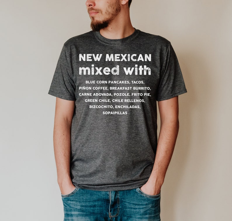 Pu&ograve; includere: Una maglietta grigio scuro con la scritta "NEW MEXICAN mixed with" e un elenco di cibi del New Mexico in bianco. L'elenco include pancake di mais blu, tacos, caff&egrave; pi&ntilde;on e altro. La maglietta &egrave; indossata da una persona.