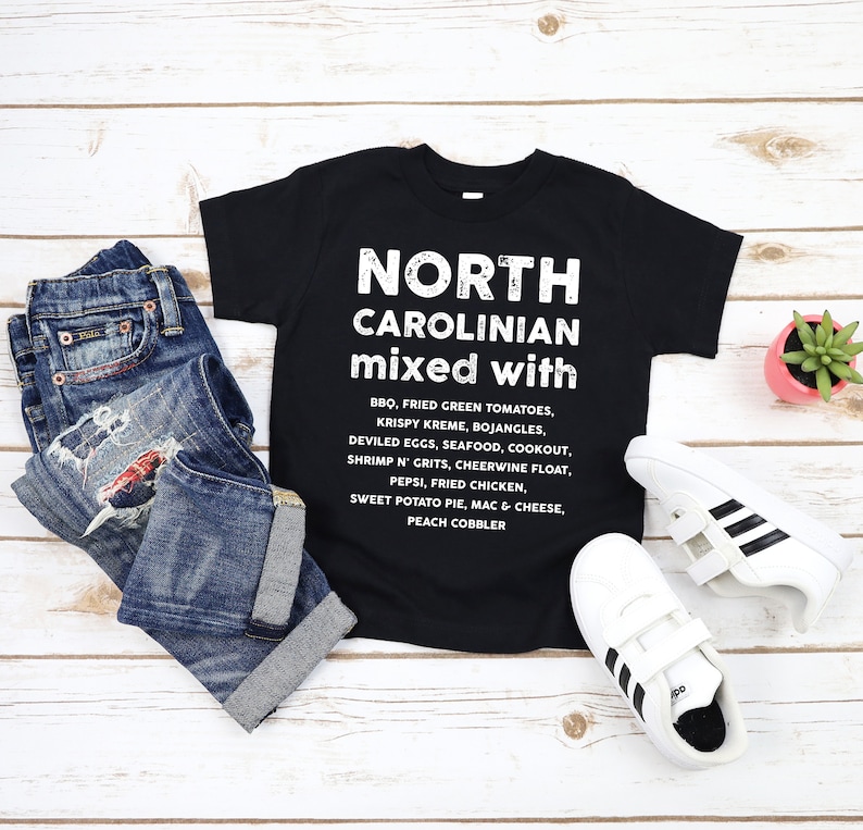May include: Black t-shirt with the text "NORTH CAROLINIAN mixed with" and a list of foods. The shirt is paired with blue jeans and white sneakers with black stripes. A small potted succulent is also present.