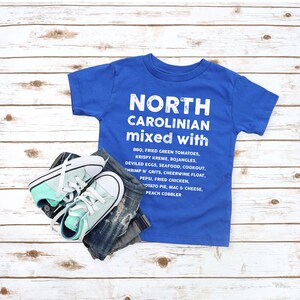 May include: A blue t-shirt with the text "NORTH CAROLINIAN mixed with" and a list of foods. The shirt is paired with denim jeans and mint green and white sneakers. The items are arranged on a light wood grain surface.