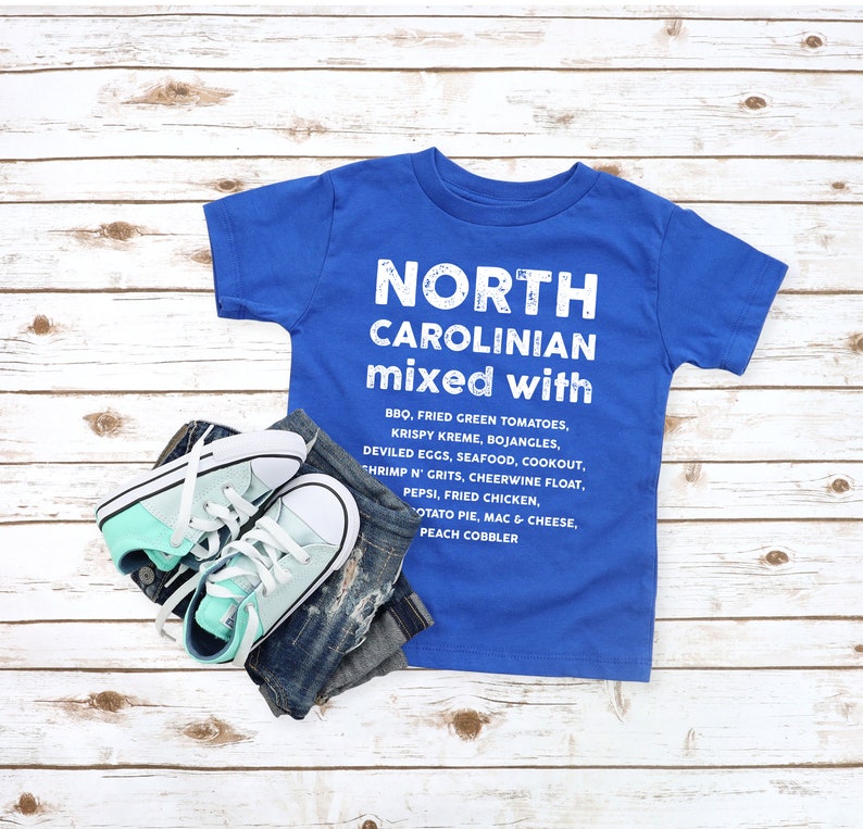 May include: A blue t-shirt with the text "NORTH CAROLINIAN mixed with" and a list of foods. The shirt is paired with denim jeans and mint green and white sneakers. The items are arranged on a light wood grain surface.
