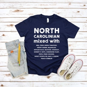 May include: Navy blue t-shirt with white text that reads "NORTH CAROLINIAN mixed with" followed by a list of foods. The shirt is paired with denim shorts, pencils, and white canvas sneakers on a wooden background.