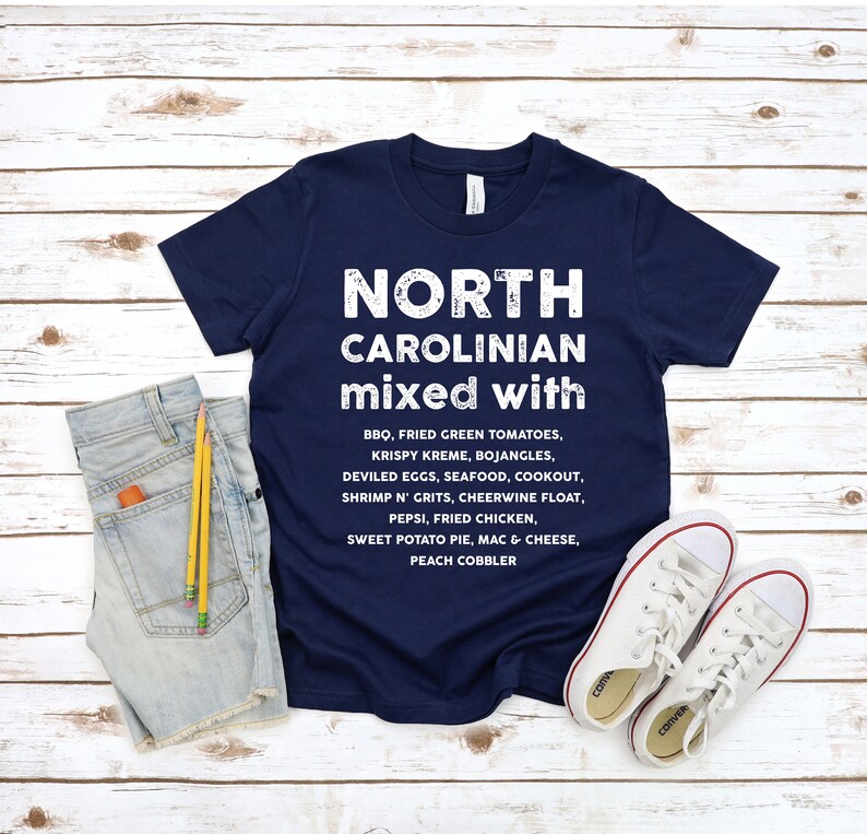 May include: Navy blue t-shirt with white text that reads "NORTH CAROLINIAN mixed with" followed by a list of foods. The shirt is paired with denim shorts, pencils, and white canvas sneakers on a wooden background.