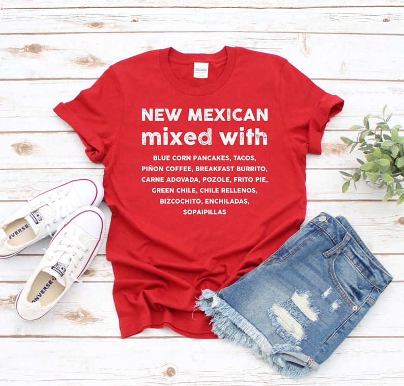 Pu&ograve; includere: Una maglietta rossa con testo bianco che elenca piatti del New Mexico come tacos ed enchiladas. La maglietta &egrave; abbinata a pantaloncini di jeans e scarpe da ginnastica bianche su una superficie di legno.