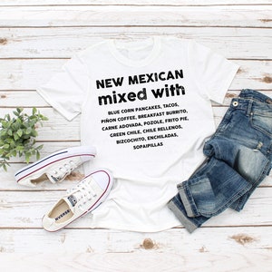 Pu&ograve; includere: T-shirt bianco con la scritta "NEW MEXICAN mixed with" e un elenco di piatti del New Mexico. La maglietta &egrave; abbinata a jeans blu e scarpe da ginnastica bianche su uno sfondo di legno chiaro.
