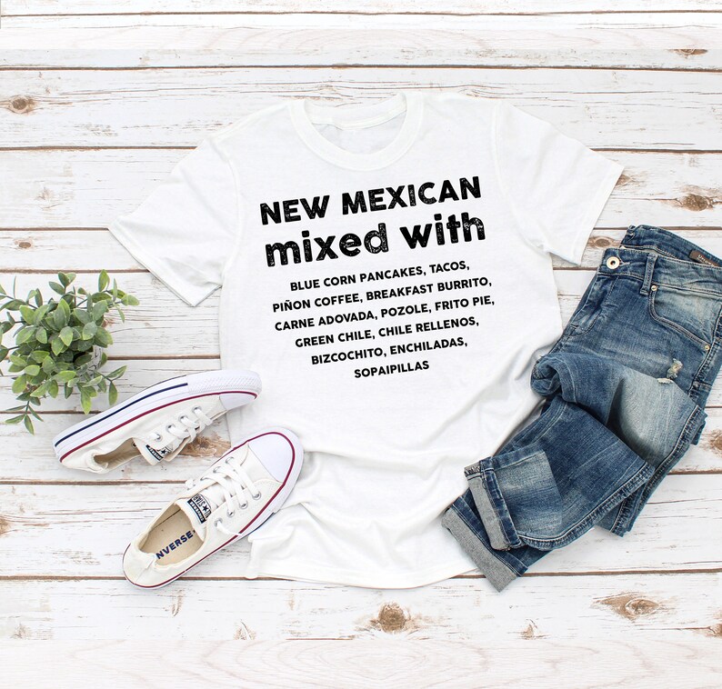 Pu&ograve; includere: T-shirt bianco con la scritta "NEW MEXICAN mixed with" e un elenco di piatti del New Mexico. La maglietta &egrave; abbinata a jeans blu e scarpe da ginnastica bianche su uno sfondo di legno chiaro.