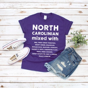 May include: A purple t-shirt with the text "NORTH CAROLINIAN mixed with" and a list of foods. The shirt is paired with denim shorts and white Converse sneakers. The shirt's text includes BBQ, fried green tomatoes, and sweet potato pie.