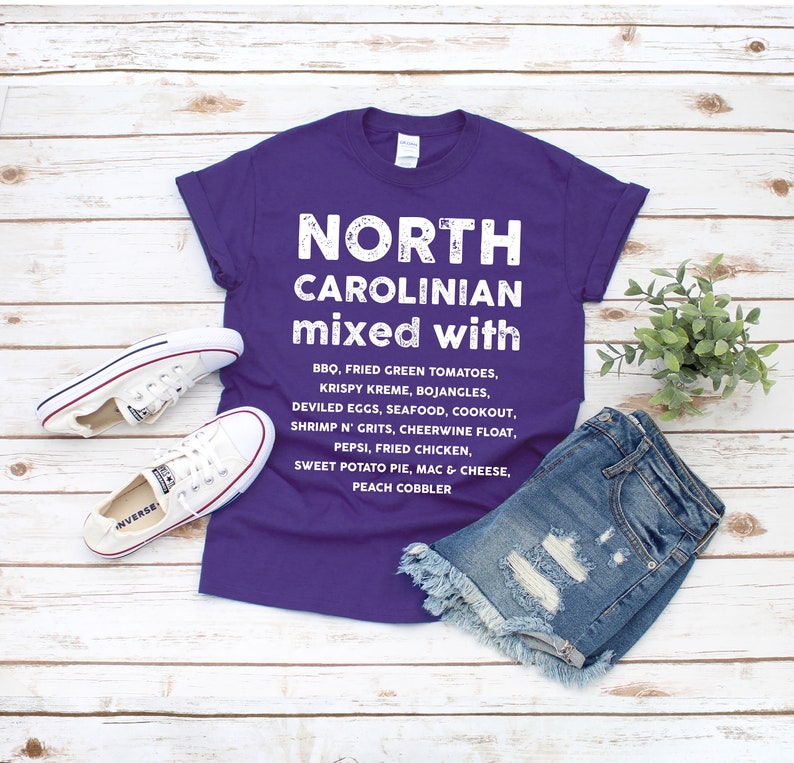 May include: A purple t-shirt with the text "NORTH CAROLINIAN mixed with" and a list of foods. The shirt is paired with denim shorts and white Converse sneakers. The shirt's text includes BBQ, fried green tomatoes, and sweet potato pie.