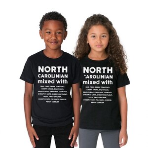 May include: Two children wearing black t-shirts with the text "NORTH CAROLINIAN mixed with" and a list of North Carolina foods in white. The list includes BBQ, fried green tomatoes, and sweet potato pie.