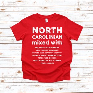 May include: A red t-shirt with white text that reads "NORTH CAROLINIAN mixed with" followed by a list of North Carolina foods, including BBQ, fried green tomatoes, and sweet potato pie. The shirt is made of a soft material.