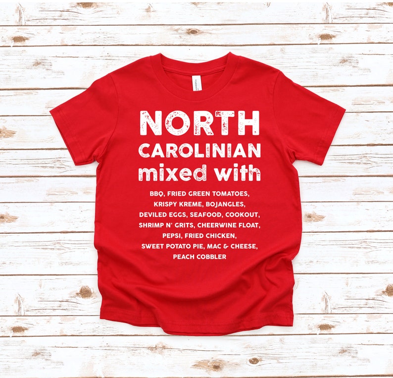 May include: A red t-shirt with white text that reads "NORTH CAROLINIAN mixed with" followed by a list of North Carolina foods, including BBQ, fried green tomatoes, and sweet potato pie. The shirt is made of a soft material.