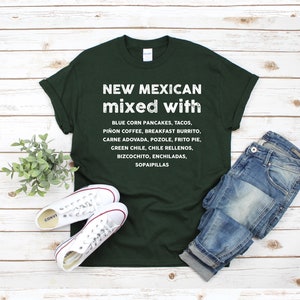 Pu&ograve; includere: T-shirt verde scuro con testo bianco che elenca piatti del New Mexico come tacos ed enchiladas. La maglietta &egrave; abbinata a scarpe da ginnastica bianche e jeans blu su uno sfondo di legno chiaro.