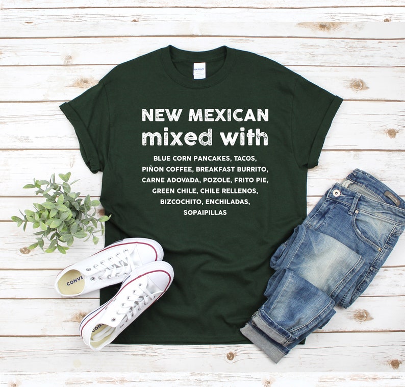 Pu&ograve; includere: T-shirt verde scuro con testo bianco che elenca piatti del New Mexico come tacos ed enchiladas. La maglietta &egrave; abbinata a scarpe da ginnastica bianche e jeans blu su uno sfondo di legno chiaro.