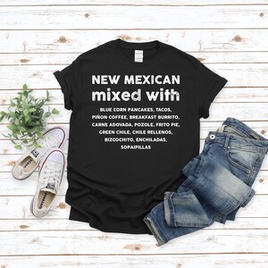 Pu&ograve; includere: T-shirt nero con testo bianco: "NEW MEXICAN mixed with" seguito da un elenco di piatti del New Mexico. La maglietta &egrave; abbinata a jeans blu e scarpe da ginnastica bianche su uno sfondo di legno chiaro.