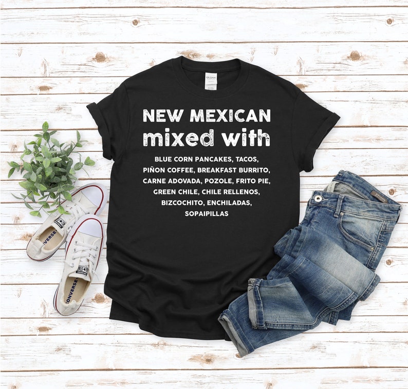 Pu&ograve; includere: T-shirt nero con testo bianco: "NEW MEXICAN mixed with" seguito da un elenco di piatti del New Mexico. La maglietta &egrave; abbinata a jeans blu e scarpe da ginnastica bianche su uno sfondo di legno chiaro.