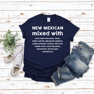 Pu&ograve; includere: Una maglietta blu navy con la scritta "NEW MEXICAN mixed with" e un elenco di cibi del New Mexico in bianco. La maglietta &egrave; abbinata a jeans blu e scarpe da ginnastica bianche su uno sfondo di legno chiaro.