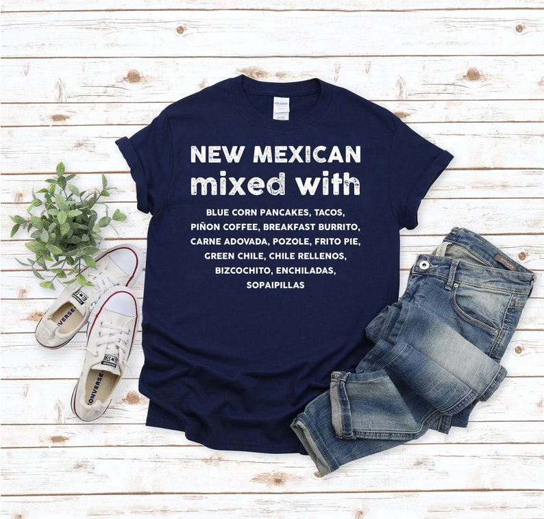 Pu&ograve; includere: Una maglietta blu navy con la scritta "NEW MEXICAN mixed with" e un elenco di cibi del New Mexico in bianco. La maglietta &egrave; abbinata a jeans blu e scarpe da ginnastica bianche su uno sfondo di legno chiaro.