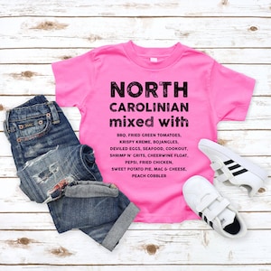 May include: A bright pink t-shirt with the words "NORTH CAROLINIAN mixed with" printed in black, along with a list of North Carolina foods. The shirt is paired with blue jeans and white sneakers with black stripes.