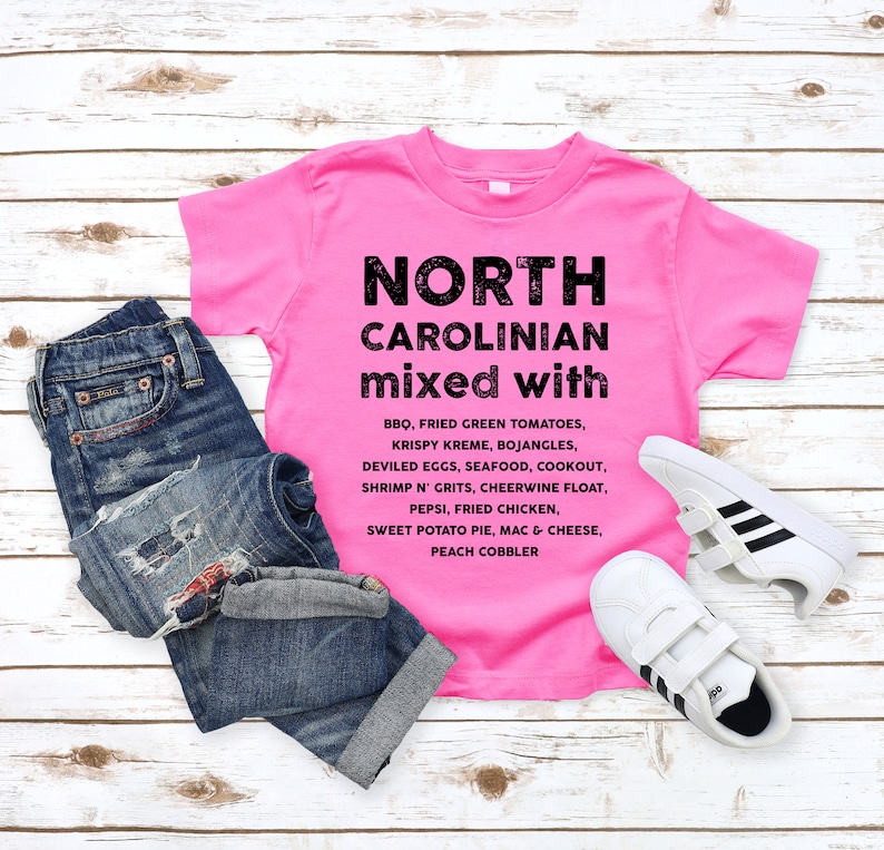 May include: A bright pink t-shirt with the words "NORTH CAROLINIAN mixed with" printed in black, along with a list of North Carolina foods. The shirt is paired with blue jeans and white sneakers with black stripes.