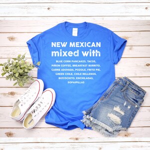 Pu&ograve; includere: Una maglietta blu con la scritta "NEW MEXICAN mixed with" e un elenco di cibi del New Mexico. La maglietta &egrave; abbinata a pantaloncini di jeans e scarpe da ginnastica bianche su una superficie di legno.