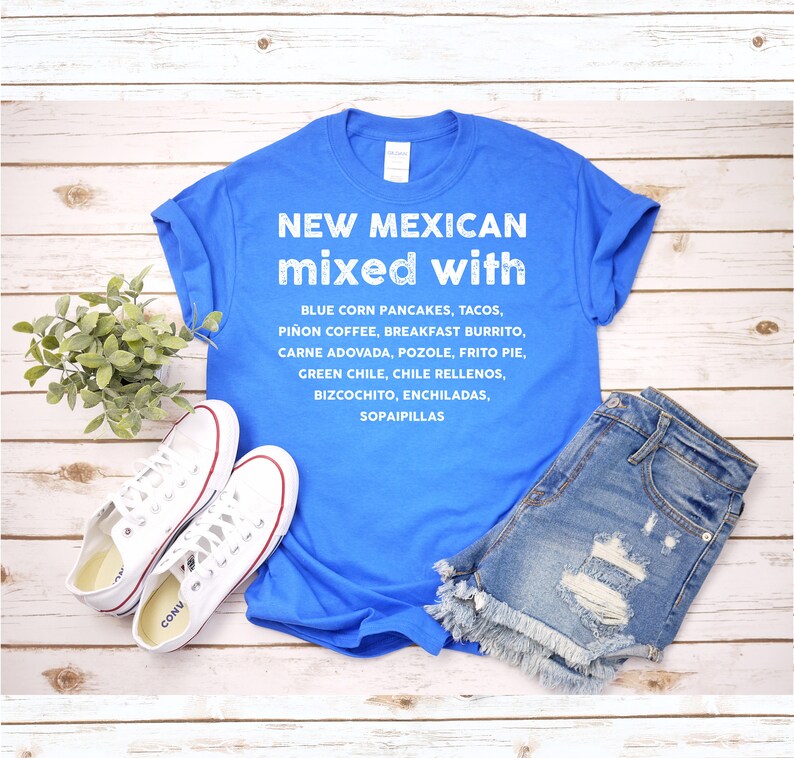 Pu&ograve; includere: Una maglietta blu con la scritta "NEW MEXICAN mixed with" e un elenco di cibi del New Mexico. La maglietta &egrave; abbinata a pantaloncini di jeans e scarpe da ginnastica bianche su una superficie di legno.