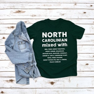 May include: A dark green t-shirt with the text "NORTH CAROLINIAN mixed with" in white. Below the text are a list of North Carolina foods. A denim jacket is draped over the shirt.