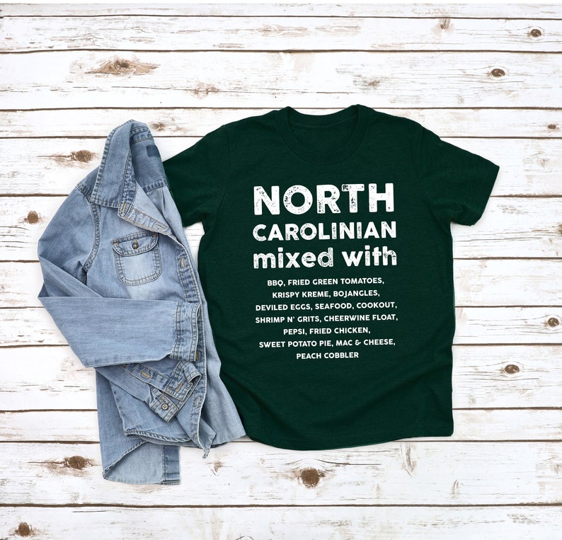 May include: A dark green t-shirt with the text "NORTH CAROLINIAN mixed with" in white. Below the text are a list of North Carolina foods. A denim jacket is draped over the shirt.
