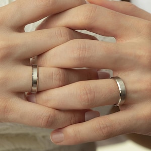 Può includere: Primo piano di due mani, una con una fede nuziale d'argento e l'altra con un anello simile. Gli anelli sono semplici, con una finitura in metallo spazzolato. Lo sfondo mostra un abito di pizzo bianco e una camicia bianca.