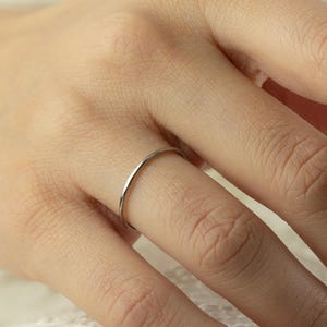Può includere: Un delicato e sottile anello d'argento è indossato su un dito. L'anello ha un design semplice e minimalista. Lo sfondo è un tessuto di pizzo bianco sporco.