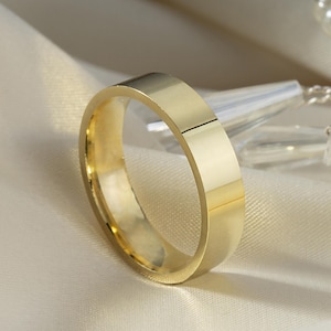 May include: A gold-colored, flat band ring. The ring is smooth and shiny, reflecting light. The ring is set against a soft, off-white fabric background, with a decorative element of pearls and crystals in the background.