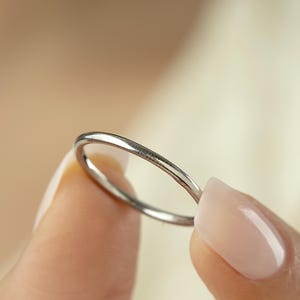 Puede incluir: Un anillo fino de plata con un acabado pulido. El anillo se sostiene entre dos dedos, mostrando su delicado diseño. El anillo es una banda simple, adecuada para el uso diario o para apilar con otros anillos. Diámetro: aproximadamente 1,5 cm.