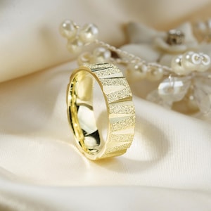May include: A gold ring with a textured, geometric design. The ring's surface features alternating smooth and textured bands, creating a striking visual effect. The ring is set against a soft, cream-colored background, highlighting its polished finish. A piece of jewelry.