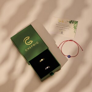 May include: A dark green sliding jewelry box with the gold logo CHEPNIQ, containing one gold-toned ring and one silver-toned ring. Beside the box is a red string bracelet with a blue evil eye charm and a thank you card with green leaf accents.
