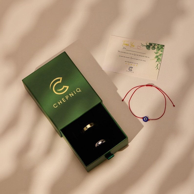 May include: A dark green sliding jewelry box with the gold logo CHEPNIQ, containing one gold-toned ring and one silver-toned ring. Beside the box is a red string bracelet with a blue evil eye charm and a thank you card with green leaf accents.
