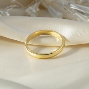 May include: A simple, polished gold band ring with a smooth, rounded surface. The ring features a classic circular design with a high-shine metallic finish, reflecting light across its curved exterior. The band is uniform in width throughout.