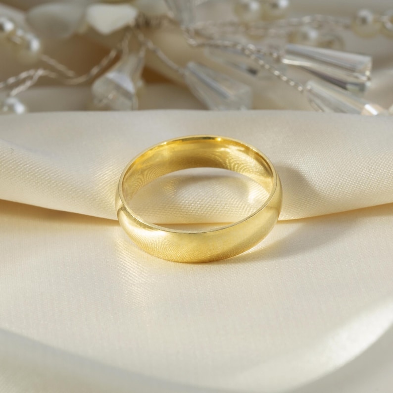 May include: A polished gold band ring with a smooth, rounded surface and a classic circular shape. The ring features a solid yellow gold construction with a high-shine finish that reflects light evenly across its entire circumference.