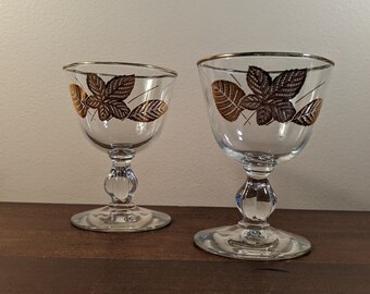 May include: Two clear glass stemmed drinking glasses with gold leaf trim and a brown leaf design.