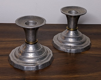 May include: Two silver candle holders with a gold band around the top of each holder. The holders have a fluted design and sit on a wooden surface.