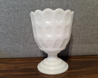 May include: White milk glass vase with a scalloped rim and a textured, raised pattern. The vase has a pedestal base.