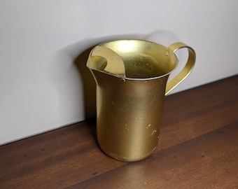 May include: A gold metal pitcher with a handle. The pitcher has a slightly dented appearance.