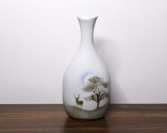 May include: White ceramic vase with a slender neck and rounded body. The vase features a painted landscape with a deer, tree, and moon. The color palette includes white, brown, and blue tones, creating a serene and natural aesthetic.