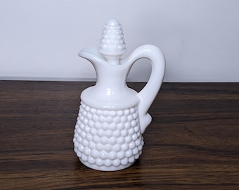 May include: A white, vintage-style creamer or small pitcher. The body has a textured, bubble-like pattern, with a curved handle and a spout. The lid is cone-shaped with a similar textured design. The creamer sits on a dark wooden surface.