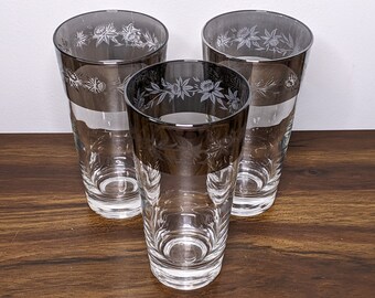 May include: Three clear glass tumblers with a silver rim and etched floral design. The glasses are on a wooden surface.