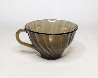 May include: A brown glass teacup with a handle. The cup has a spiral design on the exterior and a dark rim. The cup is sitting on a white surface.