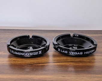 May include: Two black ashtrays with a glossy finish. One reads "Flamingo Hilton and Tower" in white text, and the other reads "Las Vegas Hilton". Both have a circular design with three notches.
