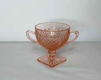 May include: A pink glass sugar bowl with two handles and a diamond pattern. The bowl sits on a square base and has a clear, open top. The glass has a vintage aesthetic.