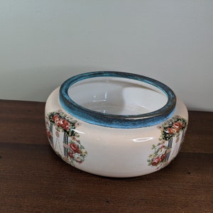 May include: A white ceramic bowl with a blue rim and floral design. The bowl has a scalloped edge and is decorated with a repeating pattern of roses and vines.